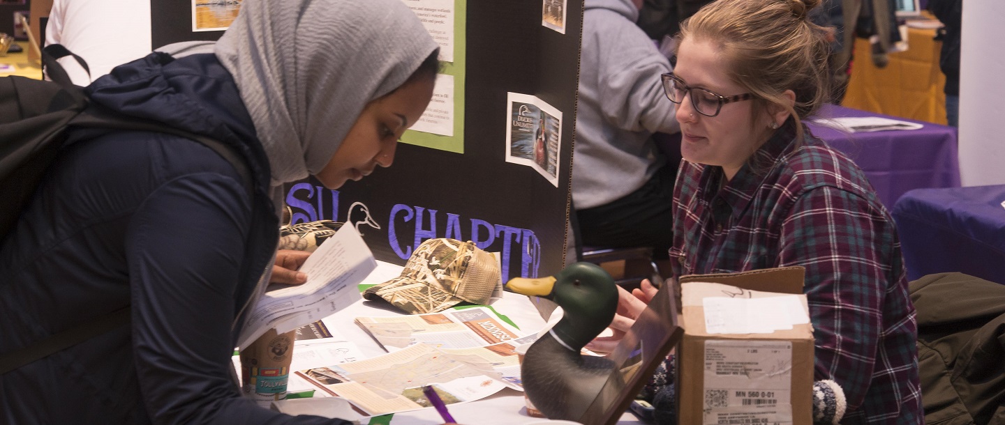 Picture of two female students, one behind table and the other across the table signing up for mailing list