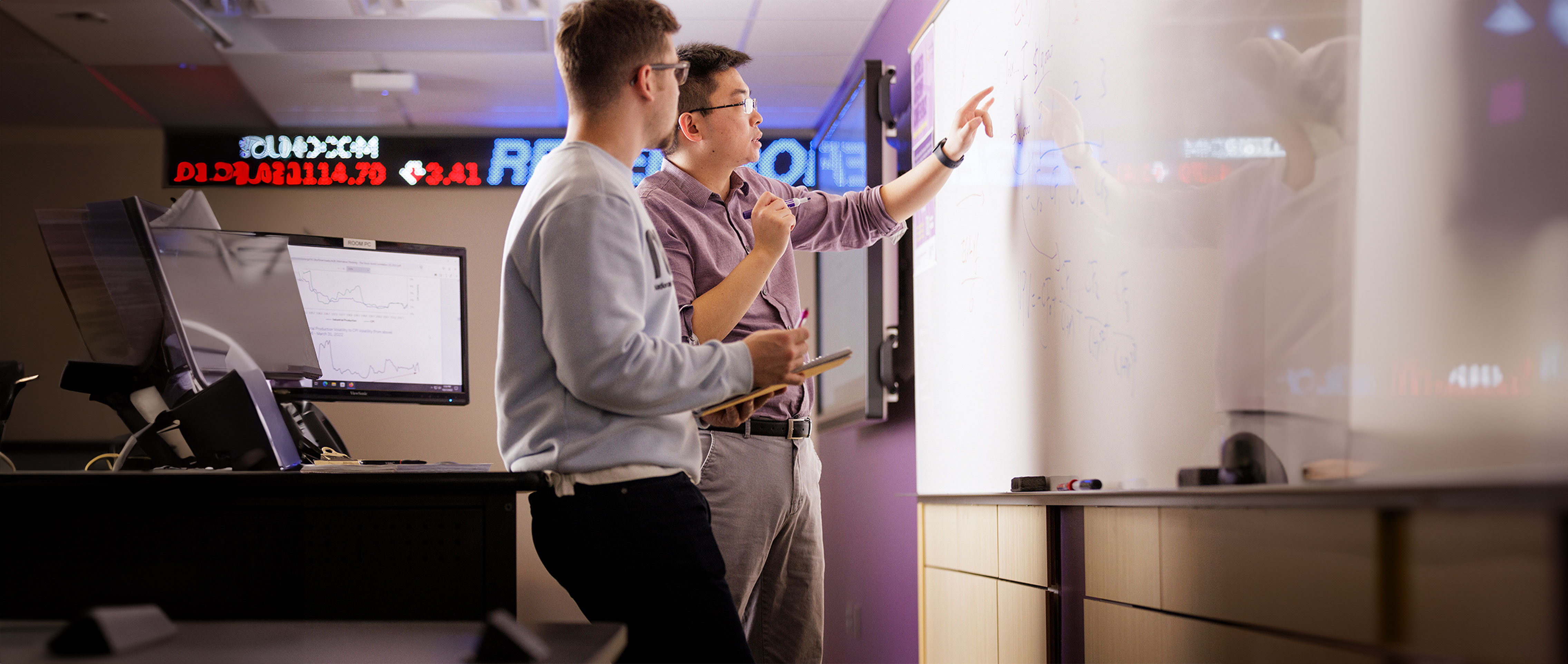 a male faculty pointing at a whiteboard and a male student is standing next to him . Both are looking at the white board 