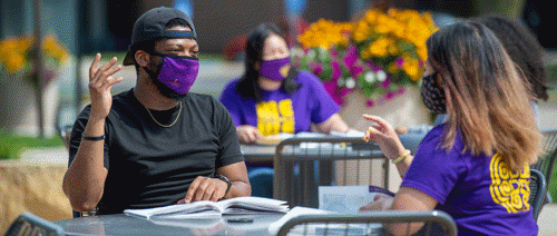 Two students with masks on sitting in one table and studying together and another student with a mask on sitting at a different table