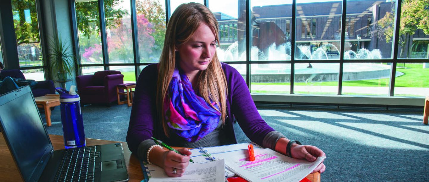 Female student studying at a desk in the library with a laptop and papers