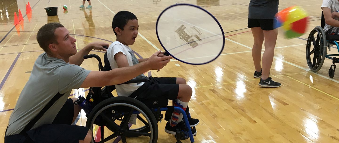 Physical Education-Developmental Adapted Physical Education students assisting kids with disabilities play in the gym with a beach ball