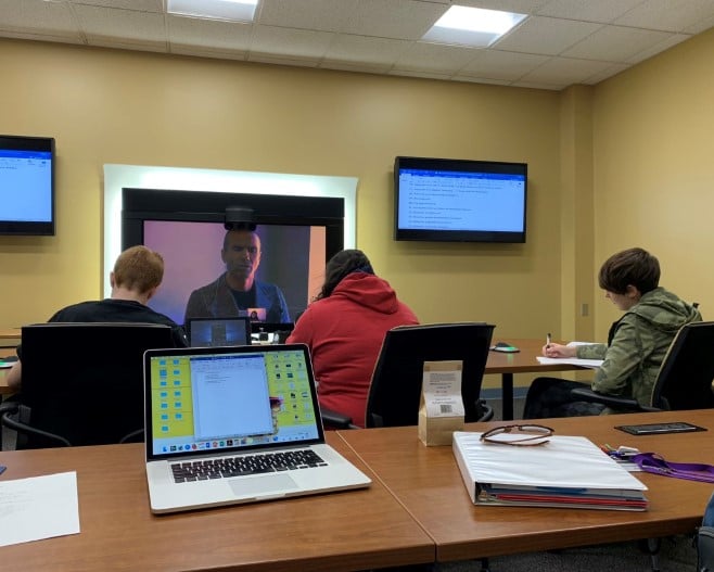 a group of people sitting at a table in a room with laptops and a large screen