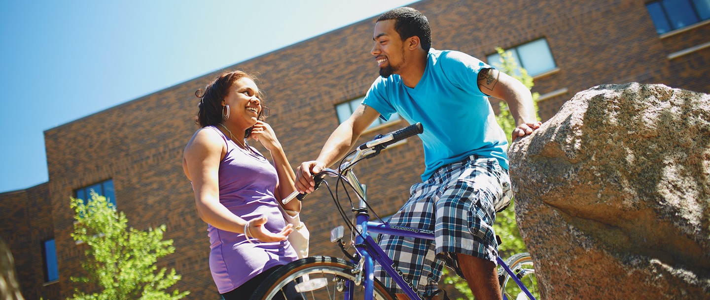 A guy sitting on a bike by a rock talking with a girl and smiling at each other