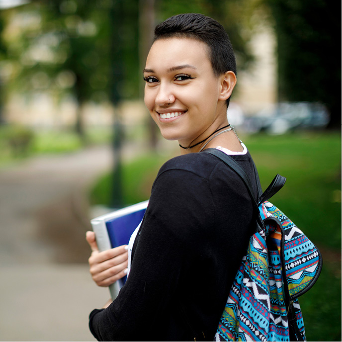 Female student wearing a backpack and holding books, smiling at the camera as she walks to class