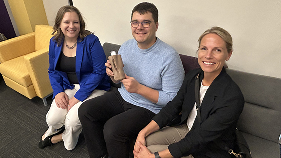 Megan Dohm, John Ruprecht and Katherine Glogowski posing on a couch as Ruprecht holds a sample of a wounded "arm" that can be created on manufacturing engineering to use in nursing