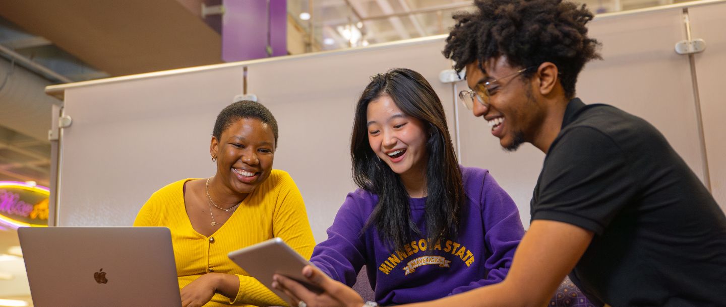 Three students sitting together and looking at a tablet and laptop, smiling.