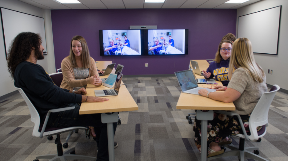 Four students sitting in a study room with laptops and two screens on the wall during a discussion at Saint Paul College