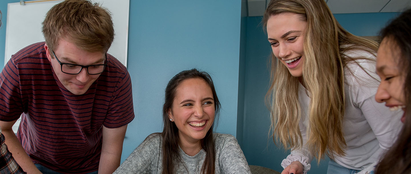 four students infront of a blue colored wall laughing while taking a look at something