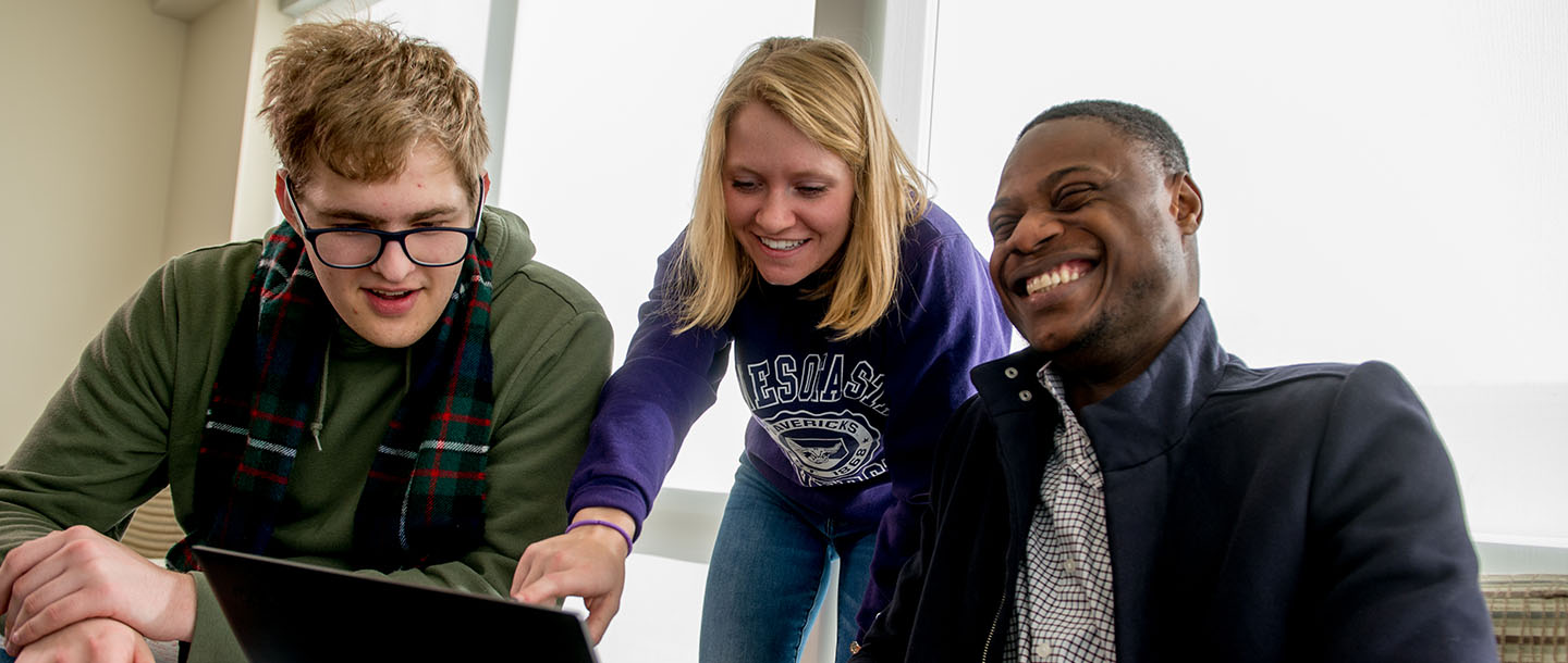 A student pointing at the screen of a computer for two other students while one student on the right is laughing