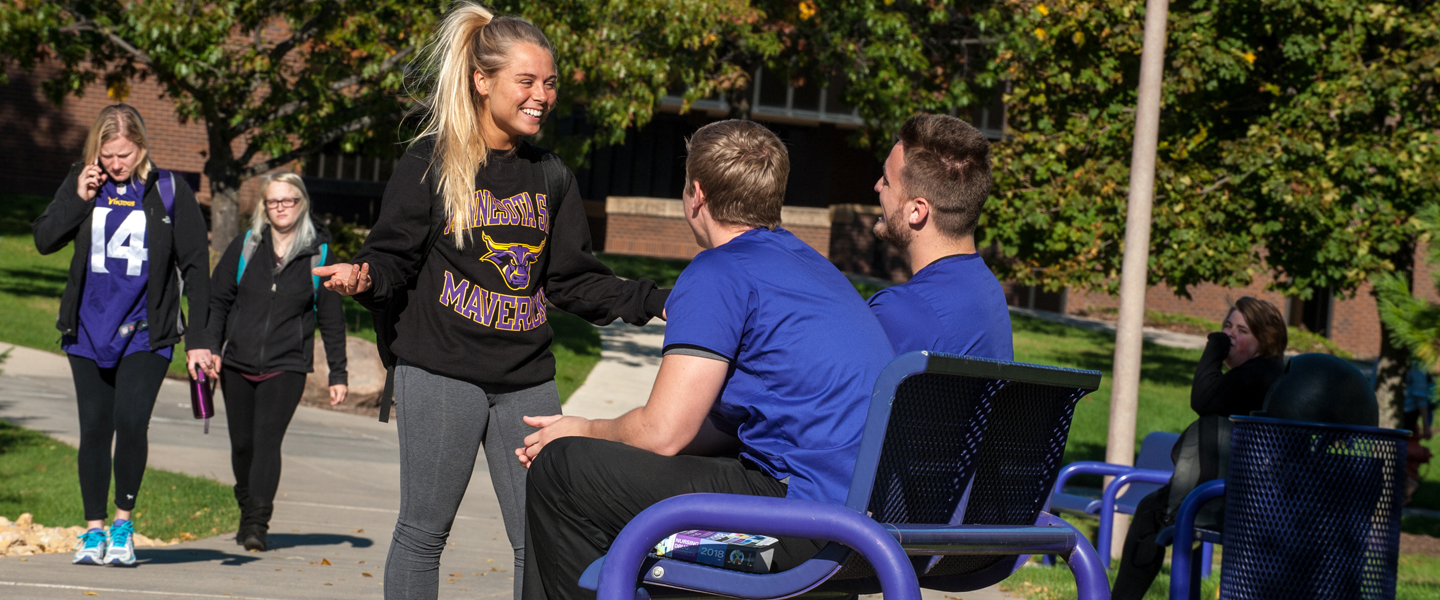 A girl talking with two boys sitting on a bench in the outside area of the college